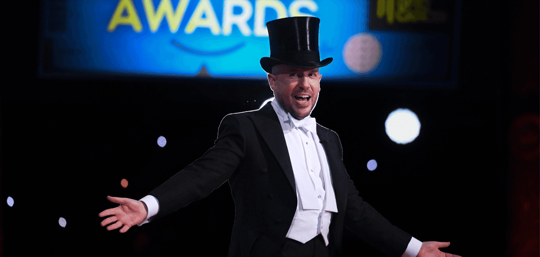 Tom Allen poses with his hands wide and smiling, wearing a black top hat and suit.