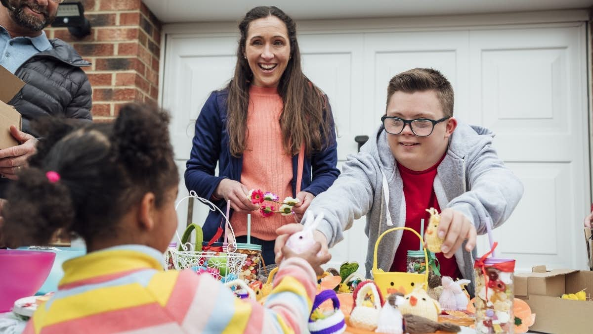 A photo of a woman and a young man behind a table at a garage sale. The young man is passing a toy to a young customer.