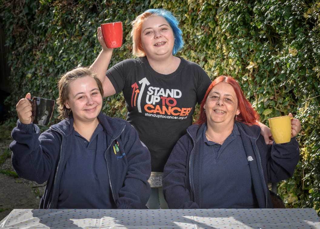 Laura and two members of her family are sitting at a table in a garden with mugs. Laura is wearing a Stand Up To Cancer branded T-shirt.