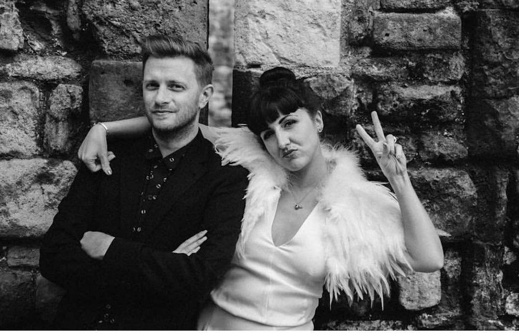 A couple on their wedding day pose for the camera. The groom is wearing a black suit, and the bride is in a white dress with feathers.