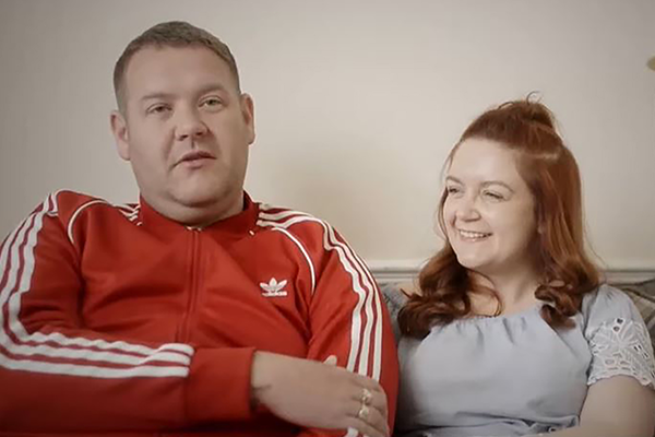 Picture of male and female. Male wearing red adidas sports jacket and woman wearning white lacey top. Both smiling and sitting on a sofa.