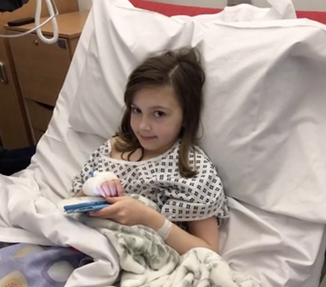 Rebecca lying in a hospital bed, phone in hand, smiling at the camera.