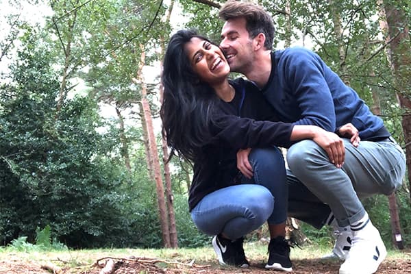 A woman and man wearing jeans crouched down smiling and embracing eachother in a wooded area.