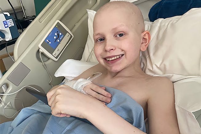 A boy with no hair on his head, smiling at the camera from his hospital bed.