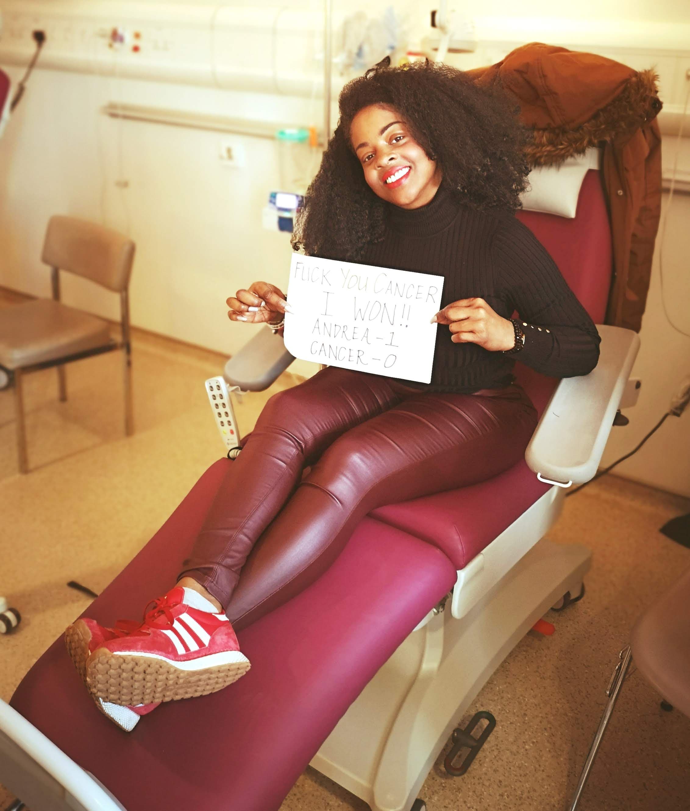 Women sitting in red medical chair holding a sign saying 'fuck you cancer, I won'.