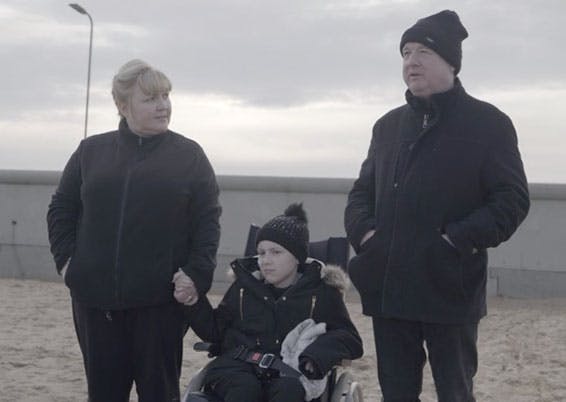 Rebecca sat in a wheelchair at the beach, holding her mum's hand and her dad stood on the other side.
