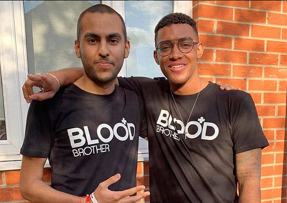 Two adult males pictured with arms around each others' shoulders. Both wearing black t-shirts with logo 'blood brothers'.