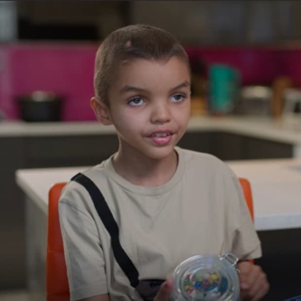 Young boy sat on orange chair wearing white t-shirt with black vertical stripe. He's holding a jar of sweets and is smiling.