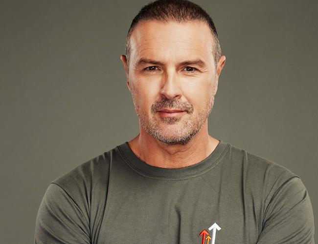 Paddy McGuinness facing the camera in a grey stand up to cancer shirt and a grey background.