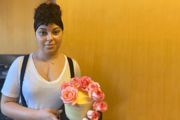 Alexe standing against a wooden background hold a cake with flowers on top.