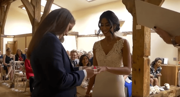 Emily putting a wedding ring on Aisha's finger with family and friends smiling behind.
