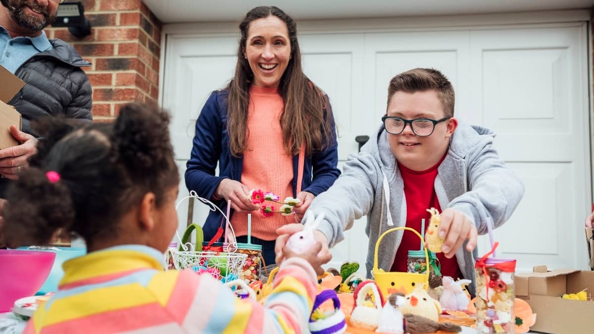 A photo of a woman and a young man behind a table at a garage sale. The young man is passing a toy to a young customer.