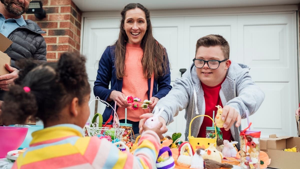 A photo of a woman and a young man behind a table at a garage sale. The young man is passing a toy to a young customer.